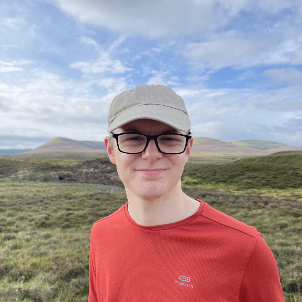 A man wearing black glasses, a beige hat and a red t-shirt. He is smiling. There are mountains, green grass and a bright sky in the background 